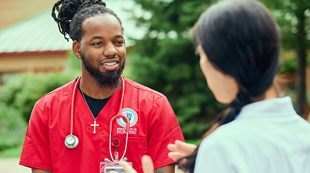 Two La Roche University students talk to each other outside on campus. One student is wearing red scrubs and a stethoscope. The other student has a white jacket.